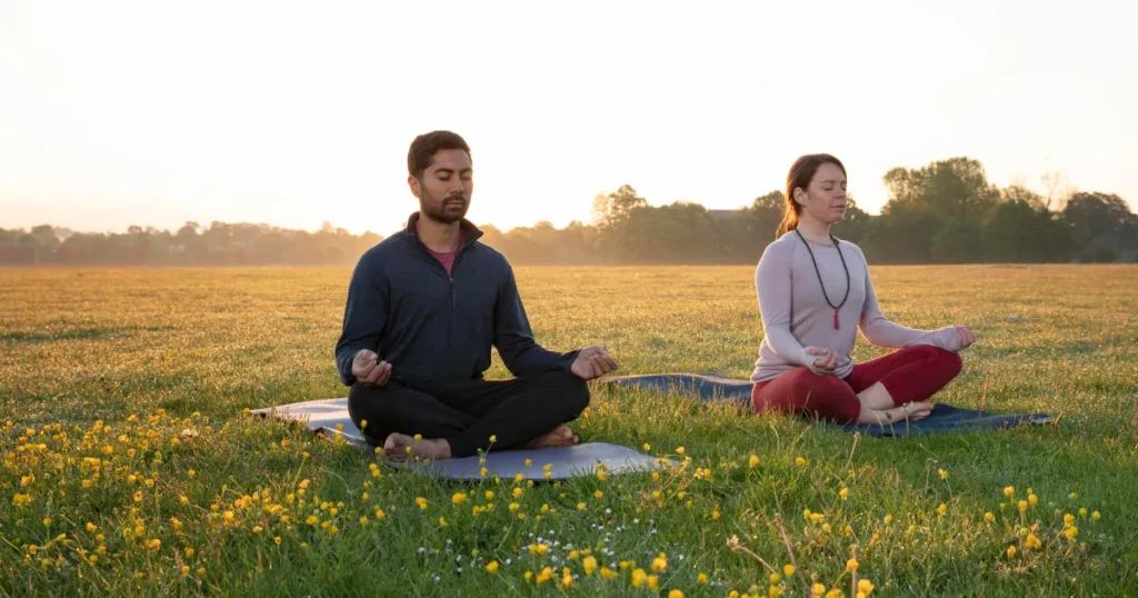 Two people sitting cross-legged on yoga mats in a field, practicing meditation at sunrise.