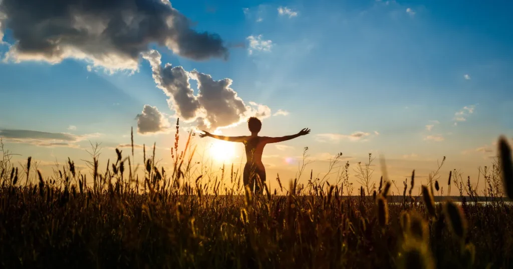 Person standing in an open field at sunrise with arms wide open, embracing light and freedom.